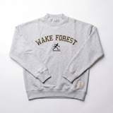 The Wake Forest Mock Neck by No Rivals is a light gray heavyweight cotton sweatshirt featuring bold black and gold WAKE FOREST lettering, a running figure graphic, DEMON DEACONS below, “NO RIVALS” on the cuffs, and a small hem tag.
