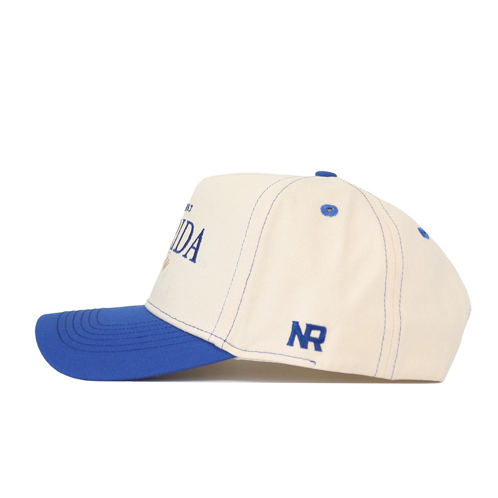 The Florida Vintage Hat is a cream-colored cap with a blue curved brim, blue stitching, and blue NR initials on the side. Made from high-quality cotton, it features matching blue eyelets and an adjustable snap back closure.