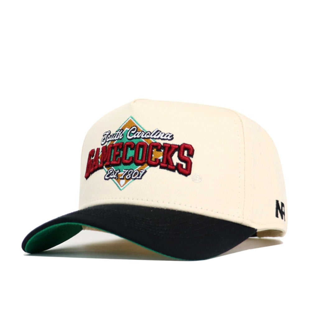 The South Carolina Ballpark Hat by No Rivals is a cream snapback made of quality cotton, featuring "South Carolina GAMECOCKS Est. 1801" embroidered in red, green, and yellow, with a structured fit, black curved brim, and green underside.