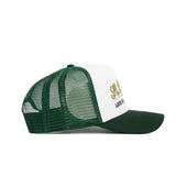 The No Rivals St.Pattys Trucker Hat features a green mesh back and bill, a white front panel with gold embroidered lettering and black text below, shown in left side profile view.