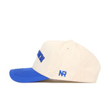The Upside Down Blue Raiders Hat by No Rivals features a cream body, royal blue brim, blue front embroidery, and NR side stitching. Made from high-quality cotton, this snapback is shown from the side against a white background.