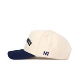 Side view of the Upside Down Kent State Hat by No Rivals in beige, featuring a navy curved brim, embroidered navy NR on the side, and a partially visible word on the front. Made from high quality cotton with a snap back closure.