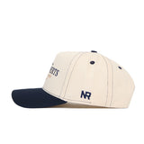 The Oral Roberts Vintage Hat by No Rivals is a beige snapback with a navy blue brim, crafted from quality cotton. It features navy stitching and "NR" embroidered in navy on the side.