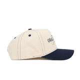The Oral Roberts Vintage Hat by No Rivals is a beige snapback made of high-quality cotton, featuring black stitching, a navy blue brim, and partially visible text on the front panel, shown from the side.