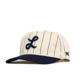 The Longwood Pinstripe Hat by No Rivals features a cream base with navy blue pinstripes, a navy brim, and an embroidered script L. It offers a snapback fit and has a small navy logo on the left side.