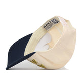 A beige and navy Michigan Wolverines Script Overlay Hat by No Rivals is flipped upside down, displaying the interior details including a QR code, “NO RIVALS” text, and part of the adjustable strap. The officially licensed cap features an embroidered logo for a premium look.